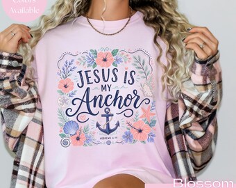 May include: A light pink crewneck sweatshirt featuring a graphic print with the text "JESUS IS MY ANCHOR" and "HEBREWS 6:19". The design includes a dark blue anchor surrounded by illustrations of pink hibiscus flowers, blue seashells, a starfish, and green foliage.
