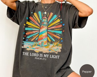 May include: Pepper-colored t-shirt featuring a colorful lighthouse graphic with the text "THE LORD IS MY LIGHT" and "PSALM 27:1". The lighthouse design includes vibrant hues of blue, yellow, orange, and green. The shirt is part of the Comfort Colors collection.