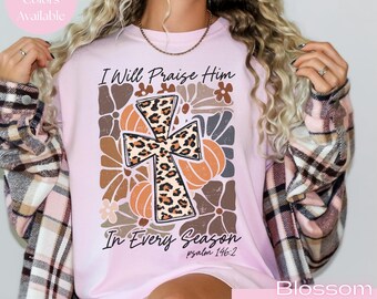 May include: A light pink t-shirt featuring a central graphic of a leopard print cross surrounded by stylized pumpkins and flowers. The text reads I Will Praise Him In Every Season and psalm 146:2 in black script. The shirt is worn under a plaid flannel.
