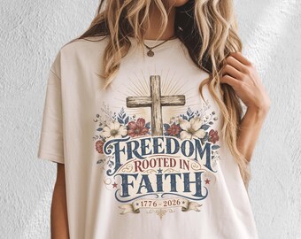 May include: A cream-colored t-shirt featuring a graphic print of a wooden cross surrounded by colorful flowers. The text reads Freedom Rooted In Faith with the years 1776-2026 below. The design includes blue, red, and white accents with star details.