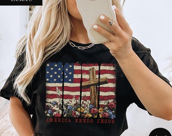 May include: Black t-shirt with an American flag design, a wooden cross, and floral details. The red text at the bottom reads "America Needs Jesus". The shirt is a comfort colors style.