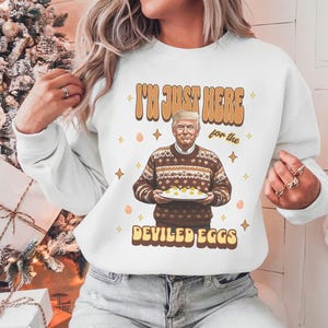 May include: White sweatshirt with a graphic of a man holding a plate of deviled eggs. The text on the sweatshirt reads "I'm just here for the deviled eggs".