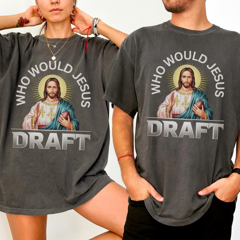 Puede incluir: Dos camisetas grises con un aspecto vintage que presentan un gr&aacute;fico de Jes&uacute;s sosteniendo un bal&oacute;n de f&uacute;tbol con el texto "Who Would Jesus Draft".