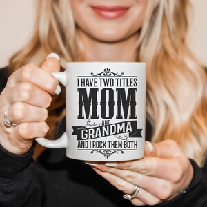 Puede incluir: Taza de cer&aacute;mica blanca con un dise&ntilde;o negro que dice "Tengo dos t&iacute;tulos Mam&aacute; y Abuela y los rockeo ambos".