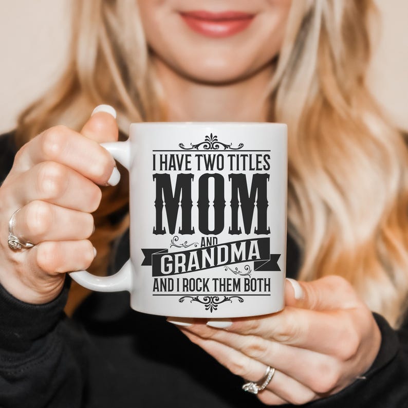 Puede incluir: Taza de cer&aacute;mica blanca con un dise&ntilde;o negro que dice "Tengo dos t&iacute;tulos Mam&aacute; y Abuela y los rockeo ambos".