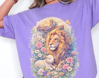 May include: A light purple t-shirt featuring a graphic print of a lion and a lamb resting together amidst a vibrant garden of pink roses and white daisies, positioned beneath a wooden cross with a small heart icon at the center of the cross.