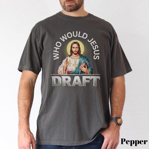 Puede incluir: Una camiseta gris oscura con un gr&aacute;fico de Jes&uacute;s sosteniendo un bal&oacute;n de f&uacute;tbol y el texto "Who Would Jesus Draft".