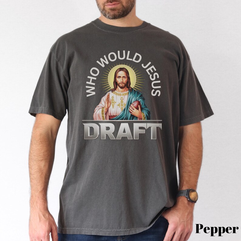 Puede incluir: Una camiseta gris oscura con un gr&aacute;fico de Jes&uacute;s sosteniendo un bal&oacute;n de f&uacute;tbol y el texto "Who Would Jesus Draft".