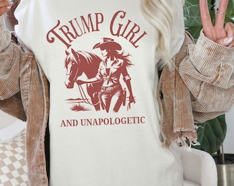May include: White t-shirt with a red graphic of a woman in a cowboy hat riding a horse. The text "Trump Girl and Unapologetic" is printed below the graphic.