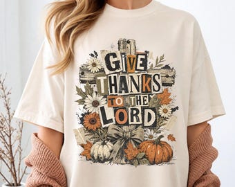 May include: A cream-colored short-sleeve t-shirt featuring a graphic print of a wooden cross decorated with autumn leaves, daisies, sunflowers, and pumpkins. The text "GIVE THANKS TO THE LORD" is printed in a bold, distressed, multi-colored font.
