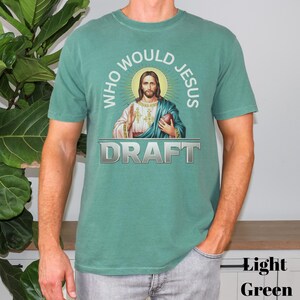 Puede incluir: Una camiseta verde claro con un gr&aacute;fico de Jes&uacute;s sosteniendo un bal&oacute;n de f&uacute;tbol. El texto "Who Would Jesus Draft" est&aacute; impreso en la camiseta.