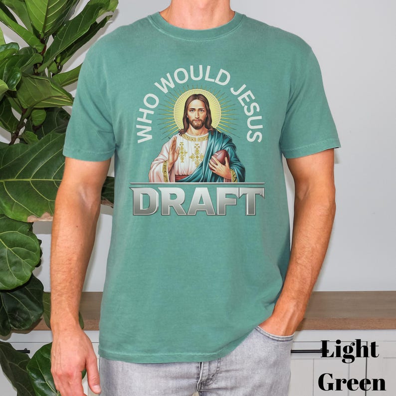 Puede incluir: Una camiseta verde claro con un gr&aacute;fico de Jes&uacute;s sosteniendo un bal&oacute;n de f&uacute;tbol. El texto "Who Would Jesus Draft" est&aacute; impreso en la camiseta.