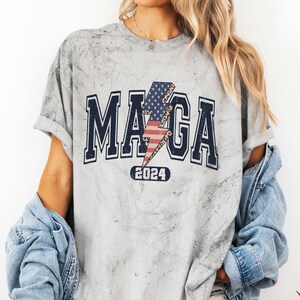 May include: A gray t-shirt with a distressed look featuring a graphic of a lightning bolt with an American flag design and the text "MAGA 2024".