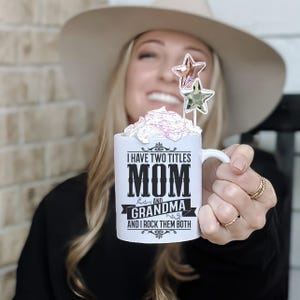 Puede incluir: Taza de cer&aacute;mica blanca con texto negro que dice "Tengo dos t&iacute;tulos Mam&aacute; y Abuela y los rockeo ambos". La taza tiene una crema batida rosa y blanca con dos adornos en forma de estrella dorada.