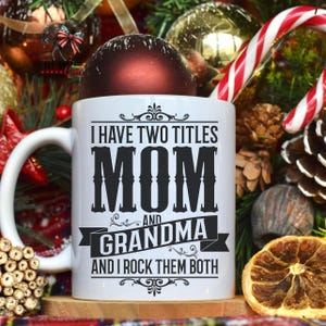 Puede incluir: Taza de cer&aacute;mica blanca con texto negro que dice "Tengo dos t&iacute;tulos Mam&aacute; y Abuela y los rockeo a ambos". La taza est&aacute; rodeada de decoraciones navide&ntilde;as, incluyendo un adorno rojo, un bast&oacute;n de caramelo y ramas de pino.