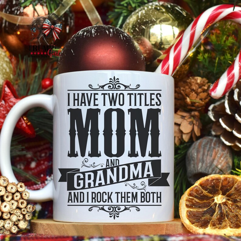 Puede incluir: Taza de cer&aacute;mica blanca con texto negro que dice "Tengo dos t&iacute;tulos Mam&aacute; y Abuela y los rockeo a ambos". La taza est&aacute; rodeada de decoraciones navide&ntilde;as, incluyendo un adorno rojo, un bast&oacute;n de caramelo y ramas de pino.