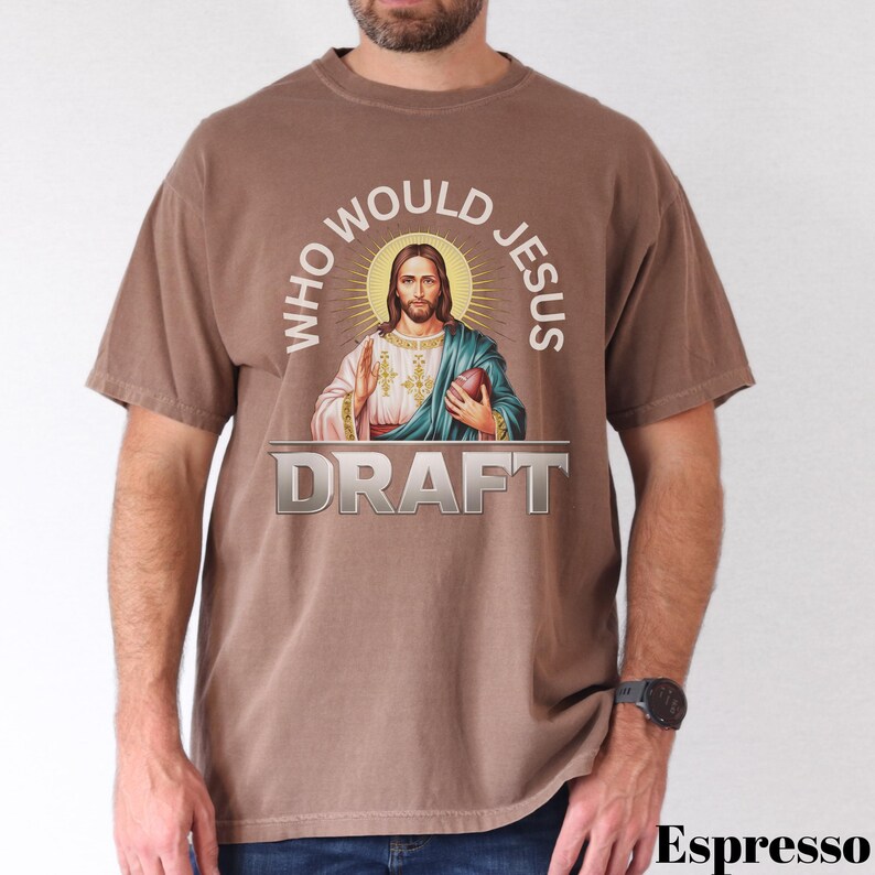 Puede incluir: Una camiseta marr&oacute;n con un gr&aacute;fico de Jes&uacute;s sosteniendo un bal&oacute;n de f&uacute;tbol. El texto "Who Would Jesus Draft" est&aacute; impreso en la camiseta.