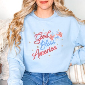 Puede incluir: Una sudadera de color azul claro con el texto "God Bless America" en letras de cuerda roja y blanca. Hay tres peque&ntilde;as banderas americanas en la esquina superior derecha del dise&ntilde;o.