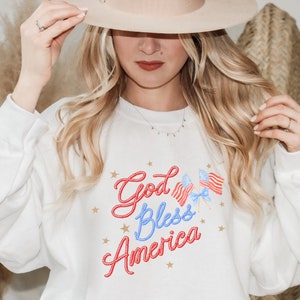 Puede incluir: Sudadera blanca con un gr&aacute;fico rojo, blanco y azul que dice "God Bless America" en una fuente tipo cuerda. El gr&aacute;fico tambi&eacute;n incluye dos peque&ntilde;as banderas americanas con un lazo azul.