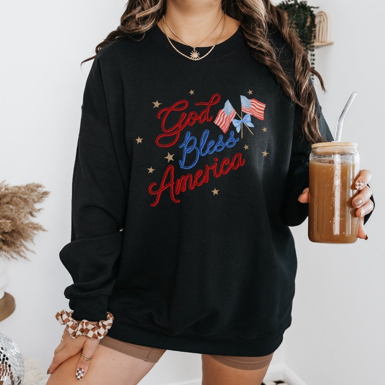 Puede incluir: Sudadera negra con un dise&ntilde;o bordado en rojo, blanco y azul que dice "God Bless America" con estrellas alrededor del texto y dos peque&ntilde;as banderas americanas.