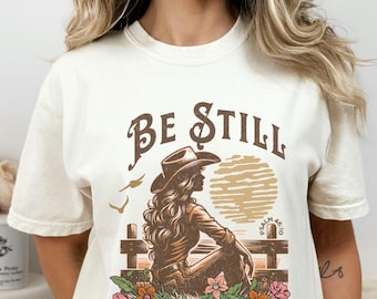 May include: White t-shirt with a brown graphic design featuring a woman in a cowboy hat sitting on a fence with flowers. The text "Be Still and Know That I Am God" is printed on the shirt. The Good Shepherd Guides Every Cowgirl.