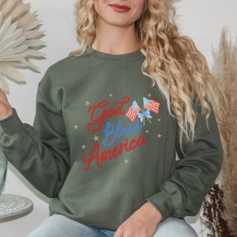 Puede incluir: Una sudadera verde con el texto "God Bless America" en letras rojas y blancas. Dos peque&ntilde;as banderas americanas est&aacute;n por encima del texto. Las banderas son rojas, blancas y azules con estrellas doradas.