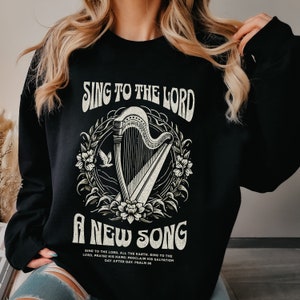 May include: Black sweatshirt featuring the text "SING TO THE LORD A NEW SONG" in a vintage style. The design includes a detailed illustration of a harp within a floral wreath, with a dove. The sweatshirt also has additional text at the bottom.
