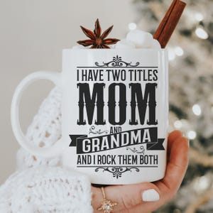 Puede incluir: Taza de cer&aacute;mica blanca con un dise&ntilde;o negro que dice "Tengo dos t&iacute;tulos Mam&aacute; y Abuela y los rockeo ambos".
