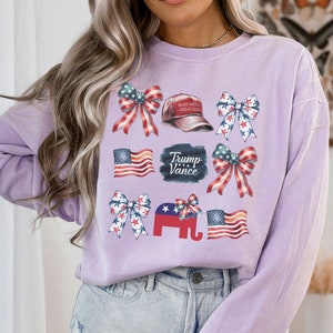 May include: A lavender long-sleeved shirt with a variety of patriotic themed graphics, including red, white, and blue bows, American flags, and a red baseball cap with the text "Make America Great Again". The shirt also features a black sign with the text "Trump Vance".