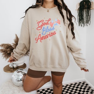 Puede incluir: Una sudadera beige con el texto "God Bless America" en letras rojas y azules con estrellas y peque&ntilde;as banderas americanas.