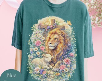May include: A blue spruce t-shirt featuring a graphic print of a lion and a lamb resting together amidst a vibrant floral arrangement. Above them is a wooden cross adorned with flowers and a small heart, all set against a soft, glowing background.