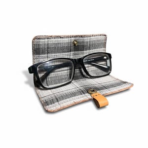 May include: Black rectangular eyeglasses rest inside a plaid patterned case. The case is open, revealing the glasses. The case has a leather tab closure. The case is gray, black, and tan.