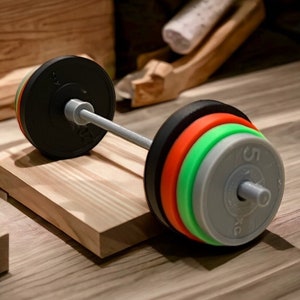 Peut inclure: Une barre d'haltères noire avec un ensemble de plaques de poids grises, vertes, orange et noires. La barre d'haltères repose sur une plateforme en bois.