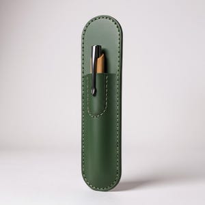 May include: A forest green leather pen case with a silver pen. The pen case is rectangular with rounded edges and a stitched border. The pen is partially visible, showing a silver clip and a wooden barrel.