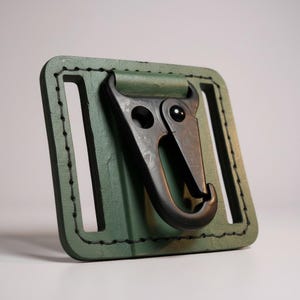 Può includere: Un passante per cintura in pelle verde con un moschettone in metallo nero. La pelle è cucita con filo nero e il moschettone è attaccato al passante con un meccanismo girevole. Il design è semplice e funzionale.