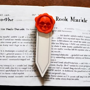 Puede incluir: Un libro abierto con un marcapáginas naranja con forma de calavera. El marcapáginas tiene una etiqueta blanca. Las páginas del libro están llenas de texto. El texto de la página dice 'MAT JAIL Bo-Oke'.