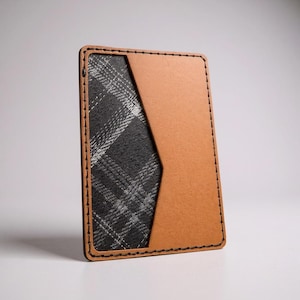 Puede incluir: Un tarjetero de cuero marrón con un bolsillo de tela a cuadros negros y grises. Los bordes están cosidos con hilo negro. El tarjetero es rectangular y está de pie.