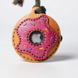 Puede incluir: Un soporte de cuero para AirTag con forma de donut, con glaseado rosa y chispas de colores. El soporte es de color marrón claro con un diseño de glaseado rosa cosido y un cordón negro para sujetarlo. El centro tiene un logotipo de Apple plateado.