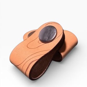 May include: A light brown leather cable organizer with a dark metal snap closure. The organizer has a curved design and etched lines, likely for decorative purposes. The item is shown against a white background.