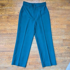 May include: Teal-colored high-waisted pants with a button closure. The pants have a straight leg design and are made of a smooth fabric. The pants are displayed on a wooden surface.
