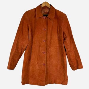 May include: A burnt orange suede jacket featuring a classic collar, long sleeves, and a front button closure with five matching buttons. The jacket has a structured fit with vertical seam detailing running down the front panels.