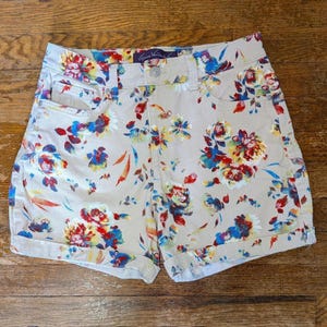 May include: Tan denim shorts with a colorful floral pattern. The shorts feature a button closure, front and back pockets, and rolled cuffs. The floral design includes red, blue, and yellow flowers.