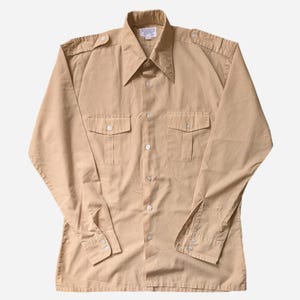 May include: A tan-colored long-sleeve shirt with two chest pockets and epaulets on the shoulders. The shirt has a pointed collar and button-down front. The cuffs have button closures. The shirt is made of a lightweight fabric.