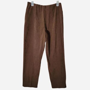 May include: Brown, straight-leg pants hanging on a white background. The pants have a simple design with a smooth texture and an elastic waistband. The pants are made of a lightweight material.