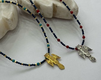 May include: Two necklaces with bird pendants. One pendant is gold-colored, the other is silver-colored. The necklaces have a dark blue beaded chain with red and turquoise accents. The pendants are detailed with intricate designs.