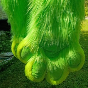 May include: A pair of bright lime green faux fur paw gloves featuring long, shaggy synthetic fibers and textured, padded paw pads in a matching shade of green. The design includes distinct toe sections with soft, plush detailing throughout.