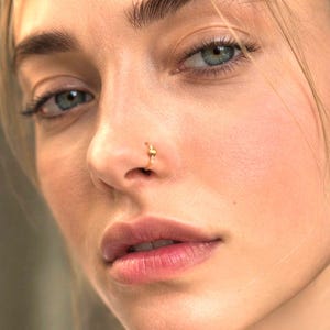 May include: A thin gold nose ring featuring a small, centred decorative element. The circular hoop is worn in the nostril, showcasing a polished metallic finish and a delicate, minimalist design that sits flush against the skin of the nose.