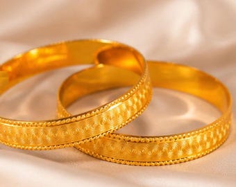 May include: Two interlocking gold bangles featuring a polished finish and a central band with a repeating textured pattern. Each bracelet has thin, beaded edges that frame the intricate, embossed design, creating a consistent metallic appearance.