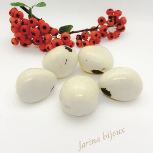 Peut inclure: Cinq perles blanches en forme d'œuf avec des marques noires. Les perles sont disposées en demi-cercle sur une surface blanche. Le texte "Jarina bijoux" est visible dans le coin inférieur droit de l'image.