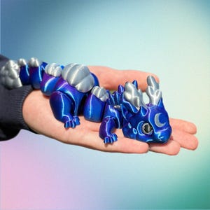 May include: A blue and silver articulated dragon figurine. The dragon has a gradient blue body, silver accents, and a crescent moon detail on its face. The dragon is a fantasy-themed collectible.